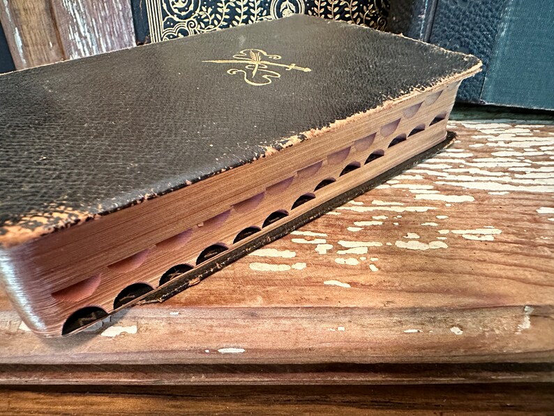 Puede incluir: Un libro vintage encuadernado en cuero negro con detalles dorados y un dise&ntilde;o decorativo en la portada. El libro est&aacute; cerrado y la encuadernaci&oacute;n es visible, mostrando que las p&aacute;ginas est&aacute;n cosidas juntas. El libro descansa sobre una superficie de madera.