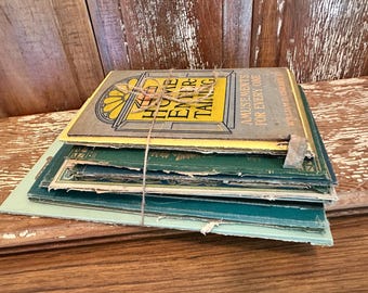 May include: A stack of vintage books tied with twine. The top book has a yellow and gray cover with the words "Home Entertaining" and "Amusements for Every One". The books are various shades of green and blue.