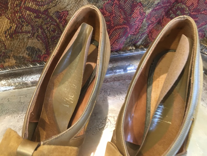 May include: A pair of brown suede shoes with a bow on the front. The shoes have a leather lining and a leather sole.