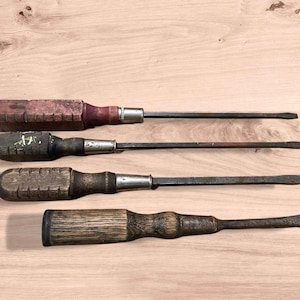 May include: Four vintage screwdrivers with wooden handles and metal shafts are arranged on a light wood surface. The handles vary in shape and color, showing signs of wear. The tools are likely used for woodworking or general repair.
