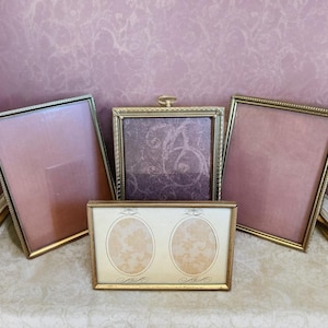 May include: A collection of vintage picture frames in various sizes and shapes. The frames have gold and silver metallic finishes, with pink and patterned backgrounds. One frame features two oval openings. The frames are arranged on a patterned surface.