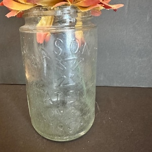 Può includere: Un vaso di vetro trasparente Mason con un'etichetta sbiadita, che contiene un finto fiore arancione e giallo. Il vaso è cilindrico con una bocca larga. La parola "MASON" è visibile.