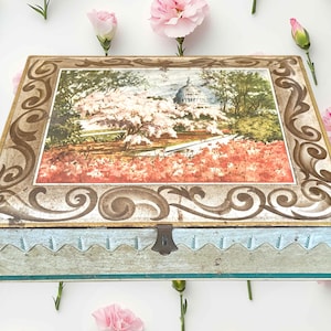 May include: A rectangular vintage metal box with a hinged lid. The lid features a framed landscape scene with a building and flowering trees. The box has a light blue base with a decorative edge and a metal clasp.