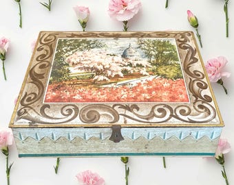 May include: A rectangular vintage metal box with a hinged lid. The lid features a framed landscape scene with a building and flowering trees. The box has a light blue base with a decorative edge and a metal clasp.
