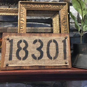 May include: A rustic wooden sign with a burlap background and the number 18301 stenciled in black paint. The sign is displayed on a wooden shelf with gold picture frames and a metal planter with a green plant.