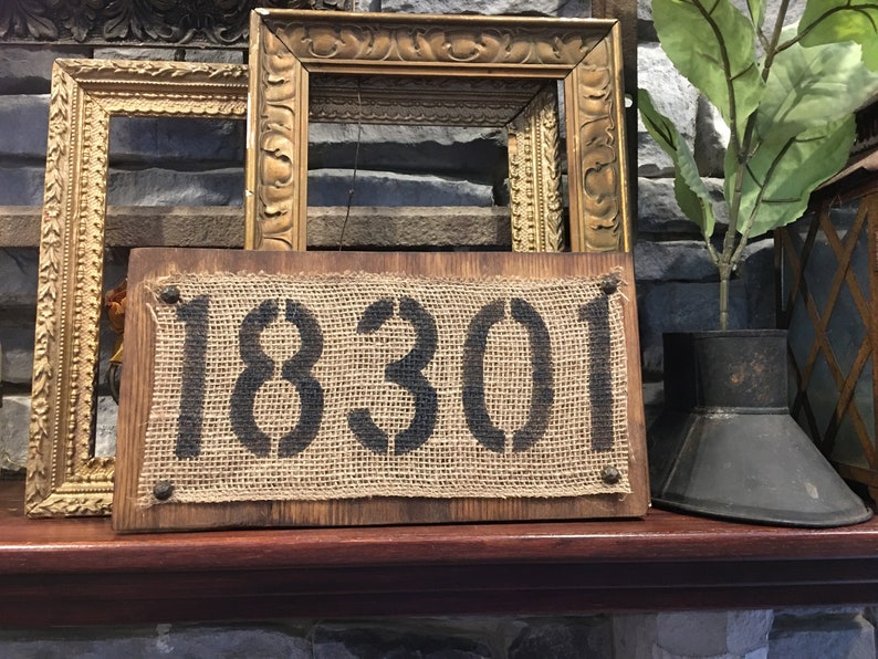 May include: A rustic wooden sign with a burlap background and the number 18301 stenciled in black paint. The sign is displayed on a wooden shelf with gold picture frames and a metal planter with a green plant.