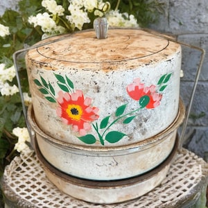 Puede incluir: Porta tartas vintage de metal blanco con tapa y asa. El porta tartas está decorado con flores rojas y amarillas pintadas a mano y hojas verdes. El porta tartas está sobre un soporte de metal blanco.