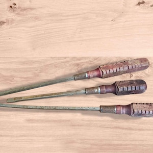 May include: Three vintage screwdrivers with wooden handles and metal shafts are arranged on a light brown wooden surface. The handles are a reddish-brown color and have a ridged design. The metal shafts show signs of age and use.