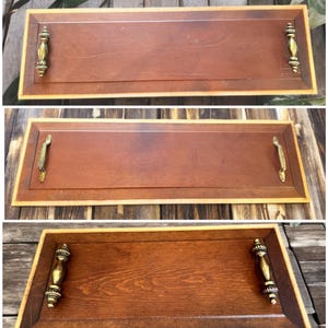 May include: A rectangular wooden serving tray with a dark brown center and a lighter brown border. It features ornate gold-colored handles on each end, adding a touch of elegance. The tray is designed for serving food or drinks.