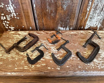 May include: An assortment of vintage C-clamps in various sizes and conditions. The clamps are made of metal and show signs of wear, with rust and discoloration. Some have orange paint. They are arranged on a weathered wooden surface.