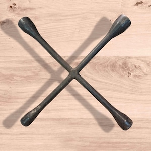 May include: A dark gray lug wrench, designed in a cross shape, rests on a light brown wooden surface. The wrench has four flared ends for gripping lug nuts. The tool is used for removing and installing wheels on vehicles.