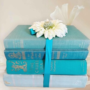 May include: A stack of four vintage books in shades of blue and teal, bound together with a teal ribbon. A large white daisy and a decorative white flower are on top. The books have gold lettering.