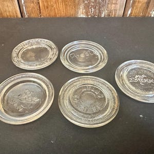 Op de afbeelding: Vijf doorzichtige glazen potdeksels met reliëftekst en ontwerpen. De deksels zijn ovaal en cirkelvormig, met het woord "Presto" en andere tekst zichtbaar. De deksels zijn gerangschikt op een donkere ondergrond.