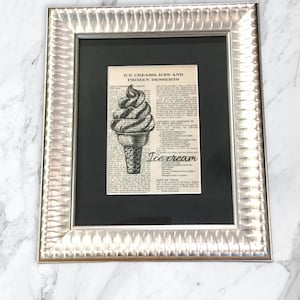 Pode incluir: Uma impressão emoldurada de um artigo de jornal vintage sobre sorvete. A impressão apresenta uma ilustração em preto e branco de um cone de sorvete com uma bola de sorvete no topo. O texto na impressão diz "Sorvetes, gelados e sobremesas congeladas" e "Sorvete".