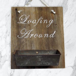 Puede incluir: Un letrero de madera rústico con el texto "Loafing Around" pintado en blanco. El letrero tiene un molde para pan de metal sujeto a la parte inferior.