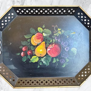 May include: Black octagonal serving tray with gold trim and handles. The tray features a hand-painted still life of fruit, including peaches, pears, cherries, and grapes. The tray's edges have a decorative lattice design.