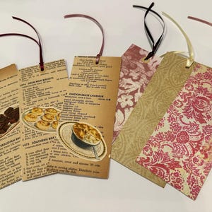 May include: A collection of handmade bookmarks. Some feature vintage recipe pages with illustrations of food, while others have patterned designs in shades of red, pink, and gold. Each bookmark has a ribbon attached.