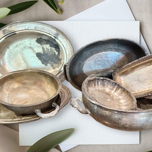 May include: An assortment of antique silver serving dishes and bowls. The collection includes an oval platter, a round bowl with handles, a rectangular tray, and several other bowls. The tarnished silver pieces are arranged on a neutral background with greenery.
