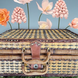 Puede incluir: Una cesta de picnic de mimbre tejido con un asa y cierre de cuero marrón. La cesta está decorada con flores de papel en tonos melocotón y rosa, con tallos verdes. El fondo es azul claro.