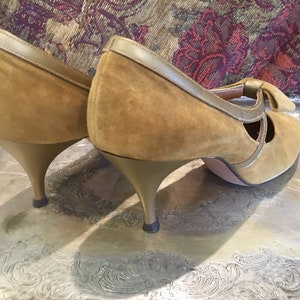 May include: A pair of brown suede heeled shoes with a bow detail on the front. The heels are gold colored.