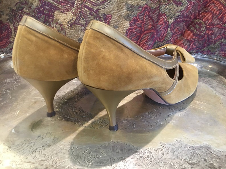 May include: A pair of brown suede heeled shoes with a bow detail on the front. The heels are gold colored.