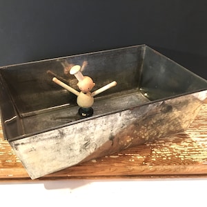 Puede incluir: Una bandeja rectangular de metal galvanizado con un acabado envejecido. Dentro de la bandeja hay una figura de chef de madera con los brazos extendidos. La bandeja está sobre una tabla de cortar de madera.