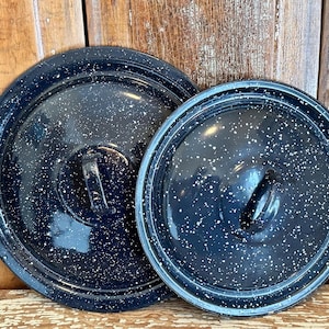 May include: Two black enamel pot lids with white speckles. The lids are round with a handle on top. The lids are different sizes and are set against a wooden background.