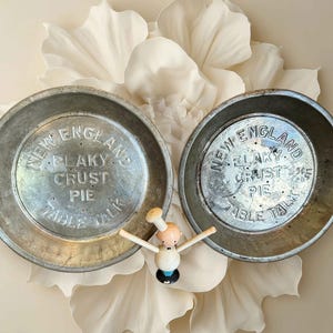 May include: Two vintage metal pie plates with the text "New England Flaky Crust Pie Table Talk" embossed on the surface. A small wooden chef figurine is positioned between the plates. The plates are set against a backdrop of white fabric flowers.