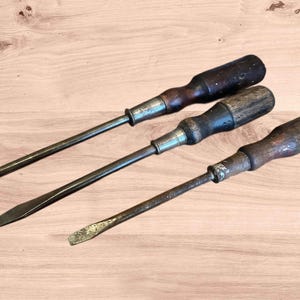 May include: Three vintage screwdrivers with wooden handles and metal shafts are arranged diagonally on a light brown wooden surface. The screwdrivers vary in size and wear, suggesting they are antique tools.