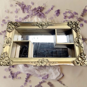 May include: A gold-painted wooden frame with ornate details, featuring a mirror and three shelves. The frame has a distressed look, with some of the gold paint chipped away.