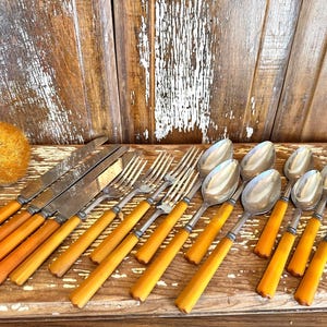 May include: A collection of vintage silverware, including knives, forks, and spoons, with orange-yellow handles and silver blades and bowls. The utensils are arranged on a wooden surface, showcasing a classic dining set.