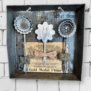 Può includere: Un vassoio in metallo vintage con tre utensili da cucina decorativi. Ogni utensile presenta un design floreale o a forma di stella. Il vassoio mostra anche una pubblicità vintage per la farina Gold Medal, aggiungendo fascino rustico.