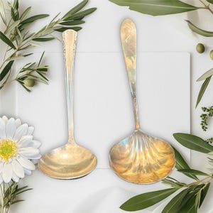 Puede incluir: Dos cucharones de servir de plata con mangos adornados. Los cucharones están sobre una superficie blanca con follaje verde.