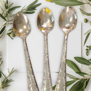 Peut inclure: Trois cuillères en argent avec des motifs ornés sur les manches. Les cuillères sont disposées en forme de triangle sur une surface blanche.