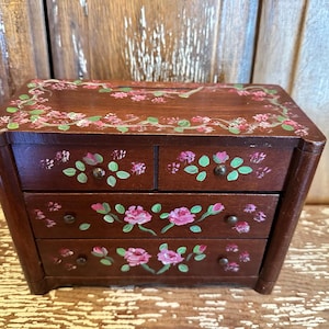 May include: A small, dark brown wooden jewelry box with four drawers. The box is hand-painted with pink roses and green leaves. The top and drawer fronts are decorated with floral designs. The box has small, round knobs on the drawers.