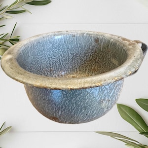 May include: An antique blue and white enamelware bowl with a speckled pattern. The bowl has a rounded shape with a wide rim and a small handle. The rim has a light brown color. The bowl is likely vintage.