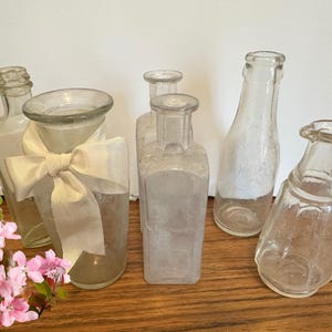 Pode incluir: Uma coleção de seis garrafas de vidro transparente com diferentes formas e tamanhos. As garrafas estão dispostas sobre uma superfície de madeira. Algumas das garrafas têm um aspeto vintage com um aspeto fosco.