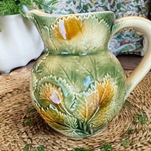 May include: A ceramic pitcher with a leaf design in shades of green, yellow, and cream. The pitcher has a curved handle and a spout for pouring. The pitcher is likely used for serving beverages.