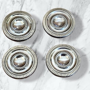 Könnte beinhalten: Vier silberfarbene, kreisförmige Schrankknöpfe. Jeder Knopf hat einen zentralen, erhabenen Kreis mit einem Perlenrand, umgeben von einem größeren, geriffelten Rand. Die Knöpfe sind auf einer weiß-grau marmorierten Oberfläche angeordnet.