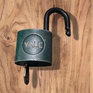 May include: An open, dark green Yale padlock with a black shackle and a key inserted. The word "YALE" is embossed on the front of the lock. The lock is set against a wooden background.