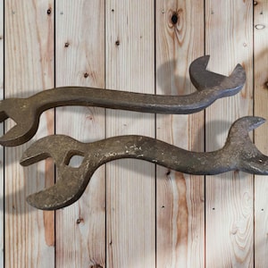 Peut inclure: Deux clés anglaises antiques et incurvées, avec des extrémités ouvertes, présentées sur un fond en bois clair. Les clés sont en métal vieilli, montrant des signes d'âge et d'utilisation. L'une des clés a un trou dans le manche.