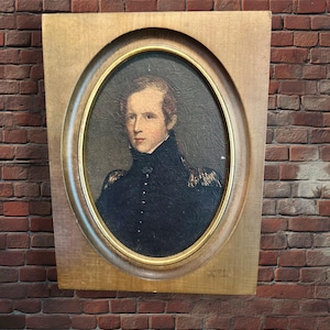 Könnte beinhalten: Ein Ölgemälde-Porträt einer Person mit hellbraunem Haar, die eine dunkelblaue, hochgeschlossene Jacke im Militärstil mit goldenen Epauletten trägt. Das Porträt befindet sich in einem ovalen Goldrahmen, der auf einer hellen Holzplatte montiert ist.