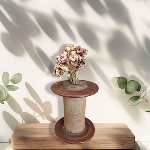Könnte beinhalten: Eine rustikale Holzspule mit getrockneten Blumen, bestehend aus einem zylindrischen Körper und zwei kreisförmigen Flanschen. Die Spule ist braun und beige, mit Blumen in Rosa- und Cremetönen. Die Anordnung steht auf einer Holzplattform.
