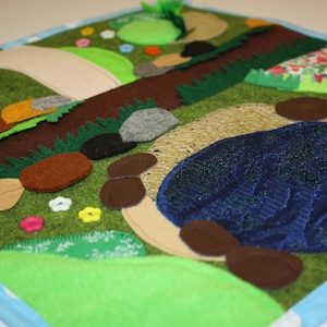 May include: A felt playmat featuring a blue pond, brown rocks, green grass, and a winding path. The playmat is ideal for imaginative play and can be used for a variety of activities.