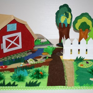 May include: A felt play set for children featuring a red barn, a white picket fence, two green trees, a brown dirt path, a blue river, and green grass.