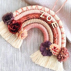 Può includere: Un arazzo da parete a forma di arcobaleno in macramè rosa e beige con un cartello con il nome in legno che dice "ZOE" e fiori di stoffa color borgogna, rosa e bianco.