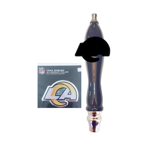 May include: A navy blue tap handle with a silver top and base. A black emblem is attached to the handle. A package with a silver and gold team emblem is visible in the background.