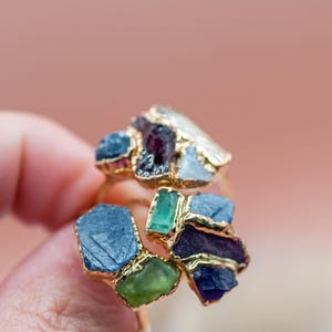 May include: A gold-toned ring featuring clusters of colorful gemstones. The ring showcases a variety of stones in shades of blue, green, purple, and white, with a gold-colored band. The gemstones are irregularly shaped, adding to the ring's unique design.