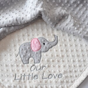 Puede incluir: Una manta de beb&eacute; suave, blanca y gris con un elefante gris bordado. El elefante tiene una oreja rosa y el texto "our little love" est&aacute; bordado debajo.