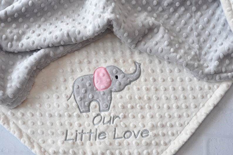 Puede incluir: Una manta de beb&eacute; suave, blanca y gris con un elefante gris bordado. El elefante tiene una oreja rosa y el texto "our little love" est&aacute; bordado debajo.
